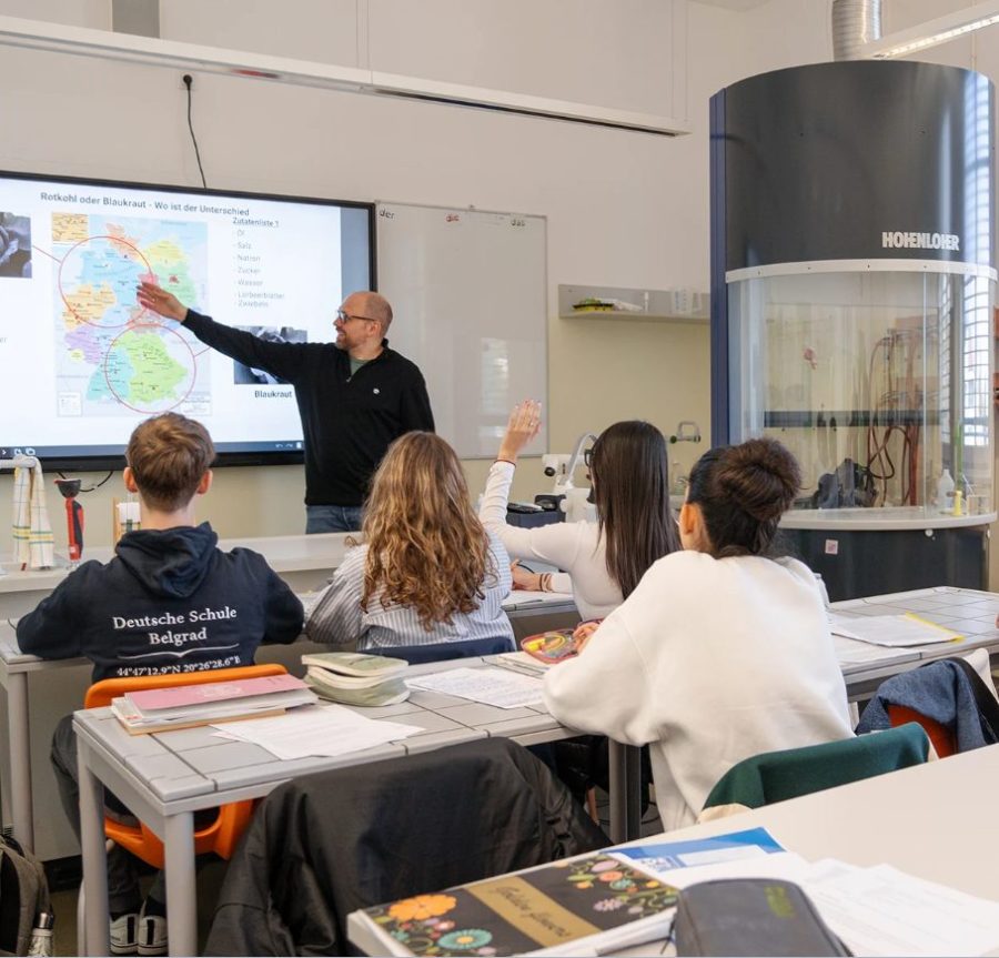 A lesson in progress: a teacher at the board with a map on screen and students at their desks, one with a hand raised
