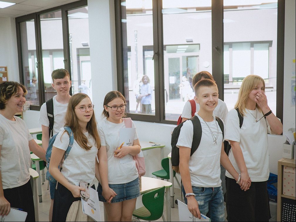 Older students in white T-shirts, smiling as they gather in a bright classroom