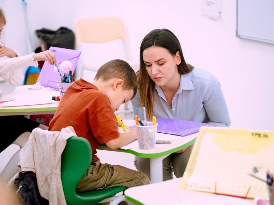 A teacher kneels beside a young pupil as he writes at his desk