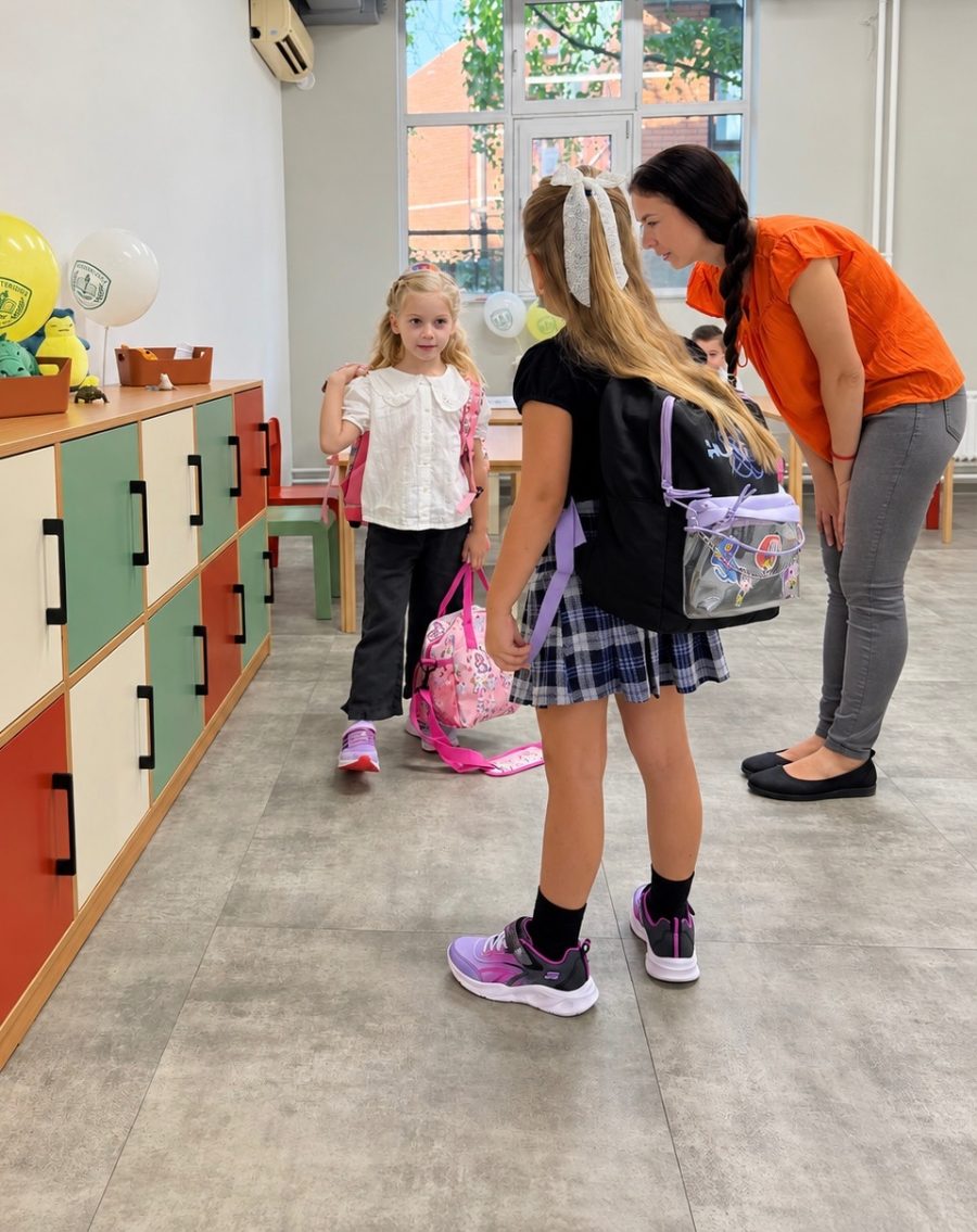 A teacher bends to greet two young pupils with their school bags beside colourful lockers