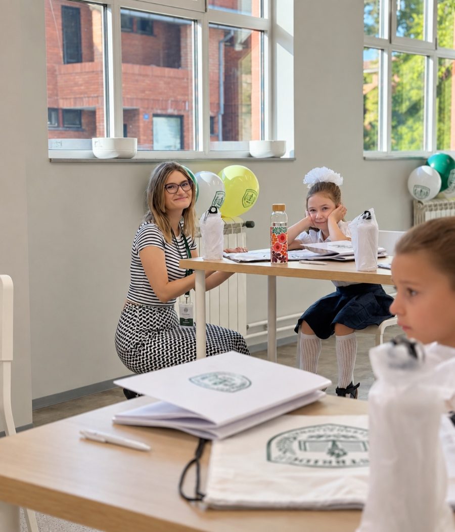 A smiling teacher sits beside a young pupil at her desk in a bright classroom